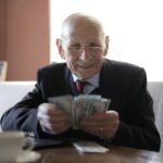 happy senior businessman counting money while sitting at table with laptop